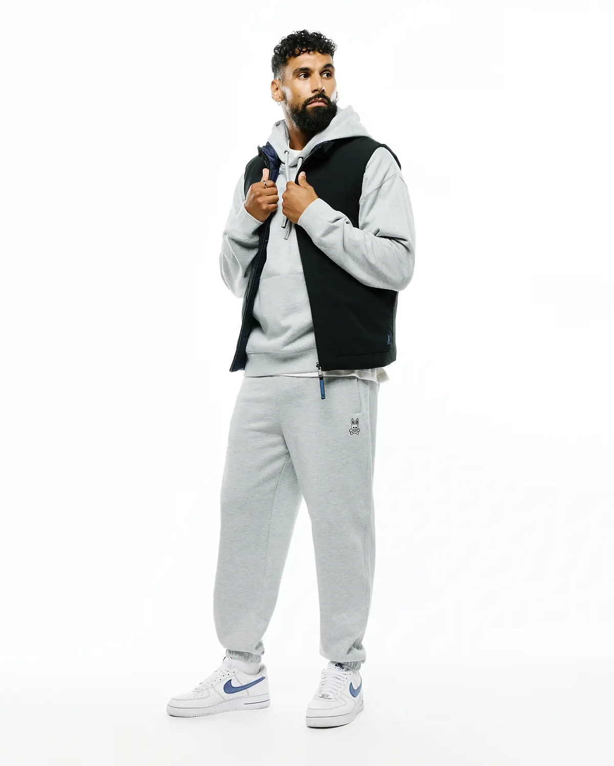 Mens Terrence Brushed Fleece Sweatpant in heather grey with a small embroidered Psycho Bunny skull logo and blue drawstring detail, paired with a matching grey relaxed-fit hoodie and a black puffer vest with navy lining.