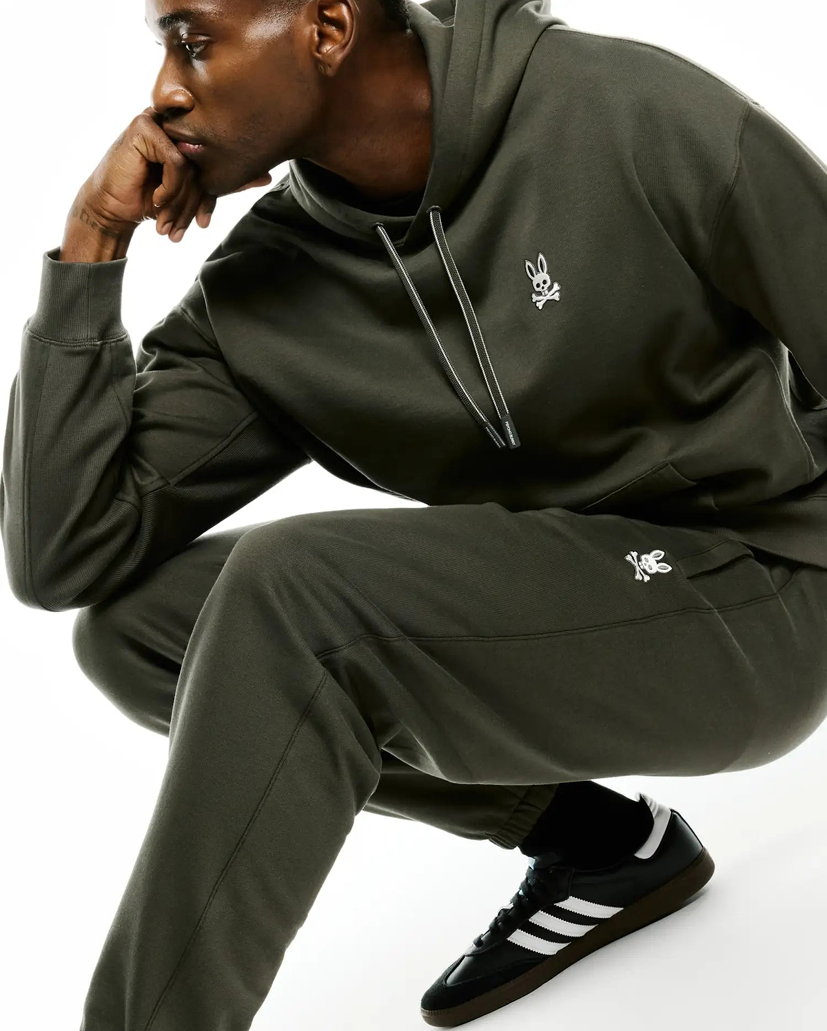 Dark olive green relaxed fit hoodie and Mens Terrence Brushed Fleece Sweatpant set, both with a small white embroidered Psycho Bunny skull logo on the left chest and hip. Hoodie features a drawstring and kangaroo pocket.