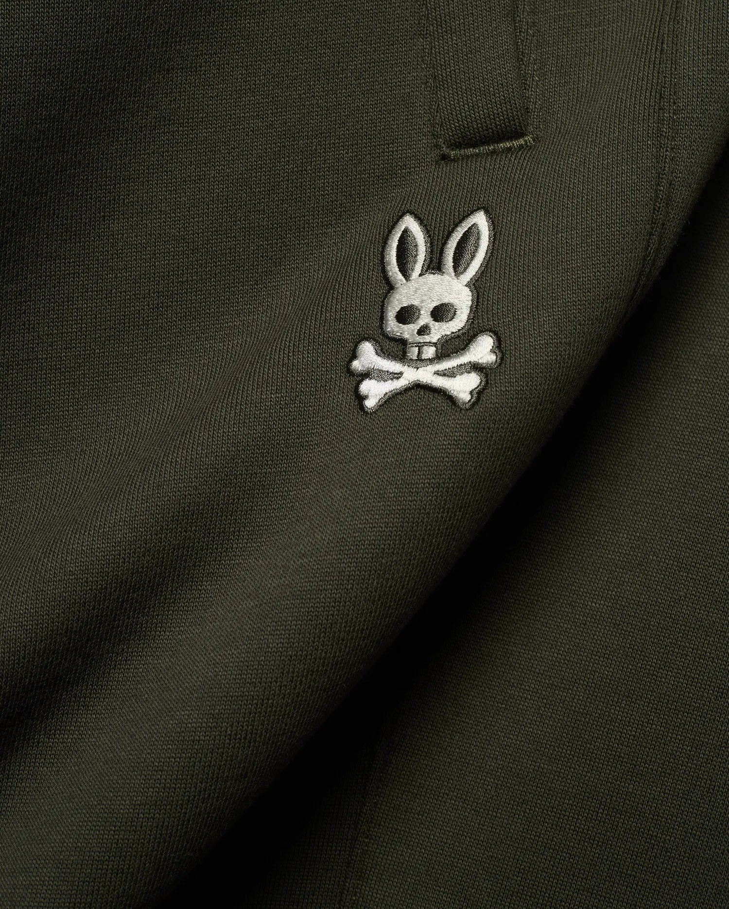 Mens Terrence Brushed Fleece Sweatpant in dark olive green, featuring a small, intricately embroidered white and silver Psycho Bunny skull logo on the upper leg, showcasing the soft brushed fleece knit texture.