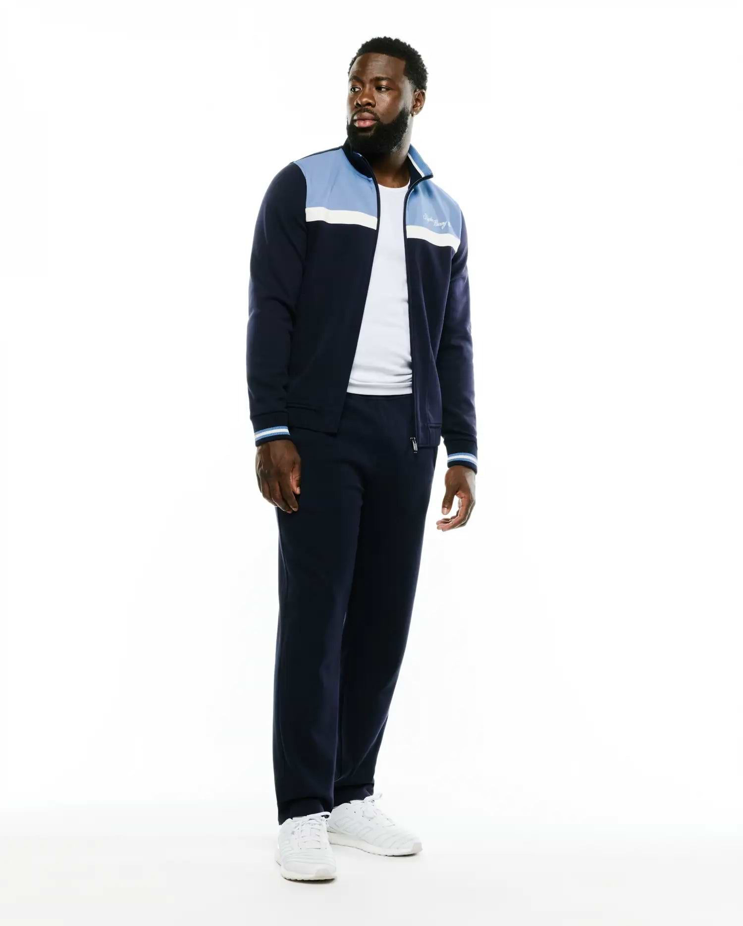 Navy blue Psycho Bunny track jacket and matching Men's Jacques Pique Track Pants. The jacket features light blue and white color-block panels, striped cuffs, and an embroidered white script logo on the chest. (1)