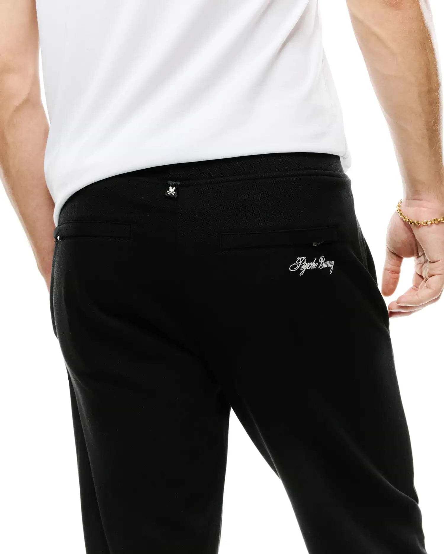 Men's Jacques Pique Track Pants in black, featuring a subtle pique texture, relaxed fit, a small white Psycho Bunny skull logo tag on the waistband, and white embroidered Psycho Bunny script on the back right pocket. (3)