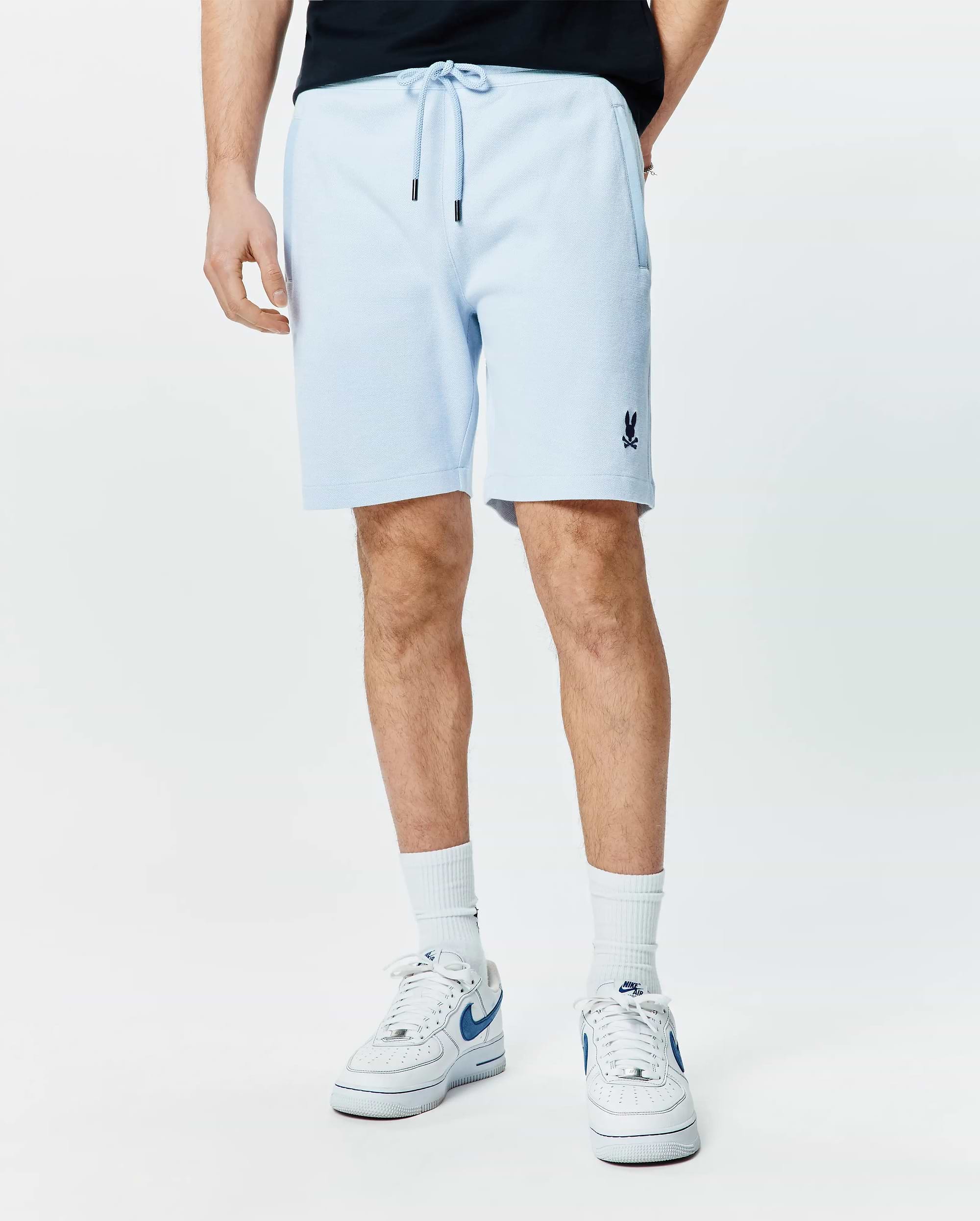 Mens Keith Short 8 light blue pique knit shorts, relaxed fit, featuring a drawstring waist with black tips, side pockets, and a small embroidered navy Psycho Bunny skull logo on the left leg.