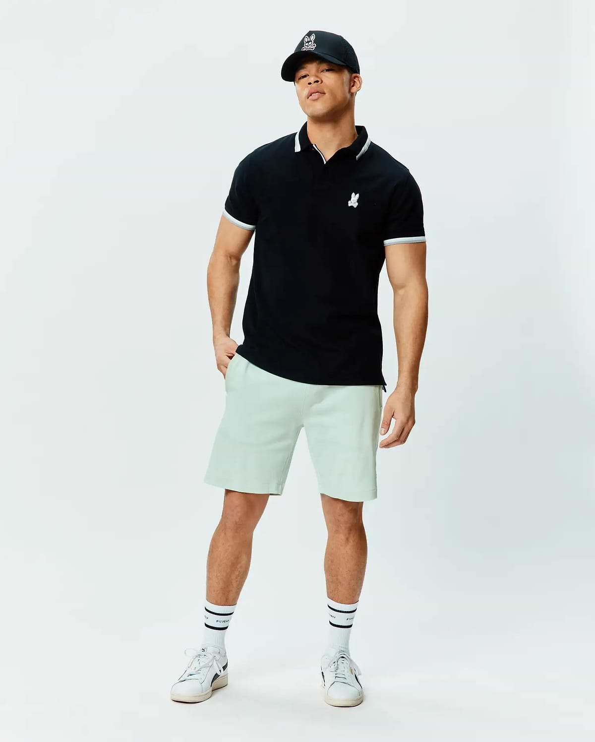 Black pique polo with white tipped collar and cuffs, featuring a small white embroidered Psycho Bunny skull logo on the chest. Paired with relaxed fit light green Mens Keith Short 8 in soft knit.