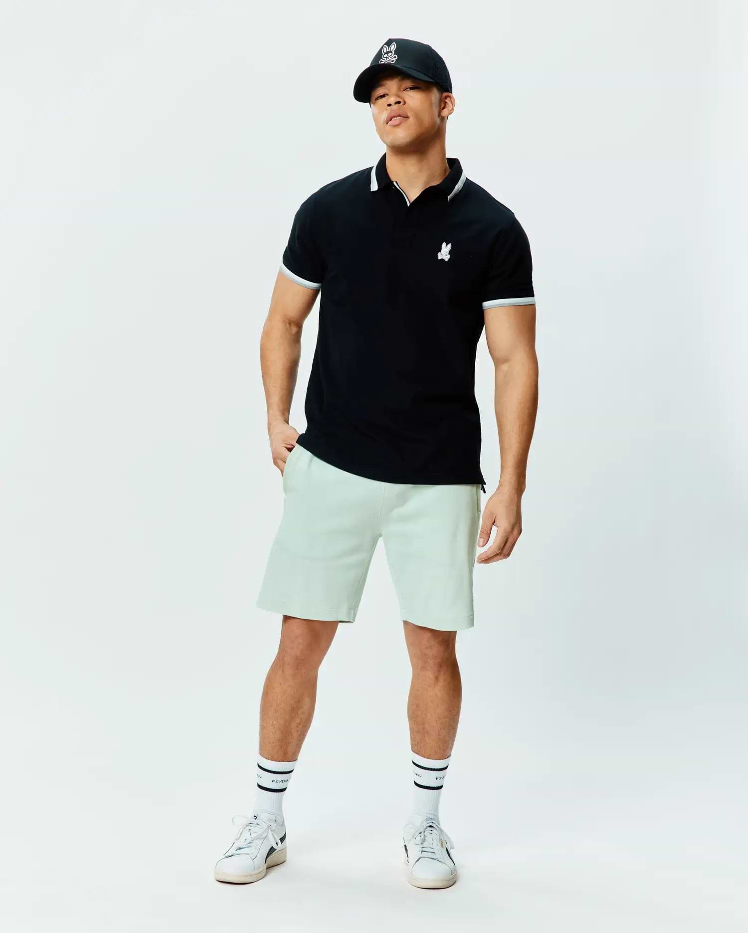 Black pique polo with white tipped collar and cuffs, featuring a small white embroidered Psycho Bunny skull logo on the chest. Paired with relaxed fit light green Mens Keith Short 8 in soft knit.