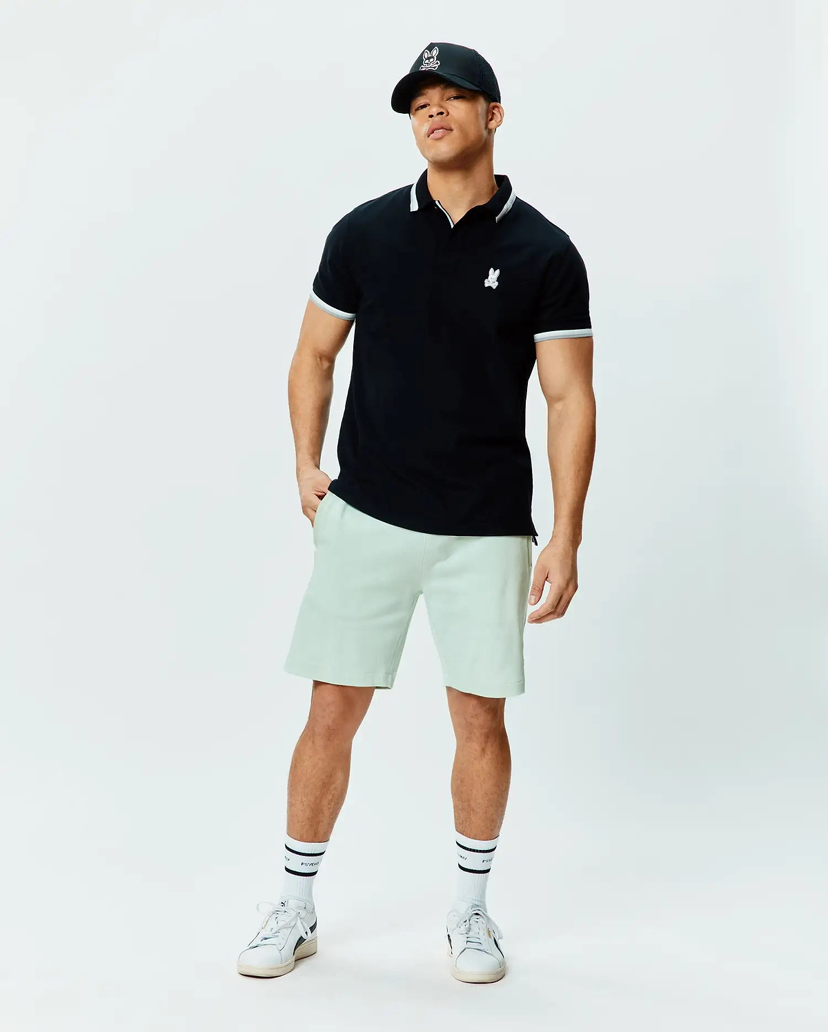 Black pique polo with white tipped collar and cuffs, featuring a small white embroidered Psycho Bunny skull logo on the chest. Paired with relaxed fit light green Mens Keith Short 8 in soft knit.