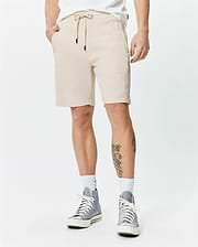 Mens Keith Short 8, sand pique knit, relaxed fit shorts with drawstring waist, black tips, and a small white embroidered Psycho Bunny skull logo on the left thigh.