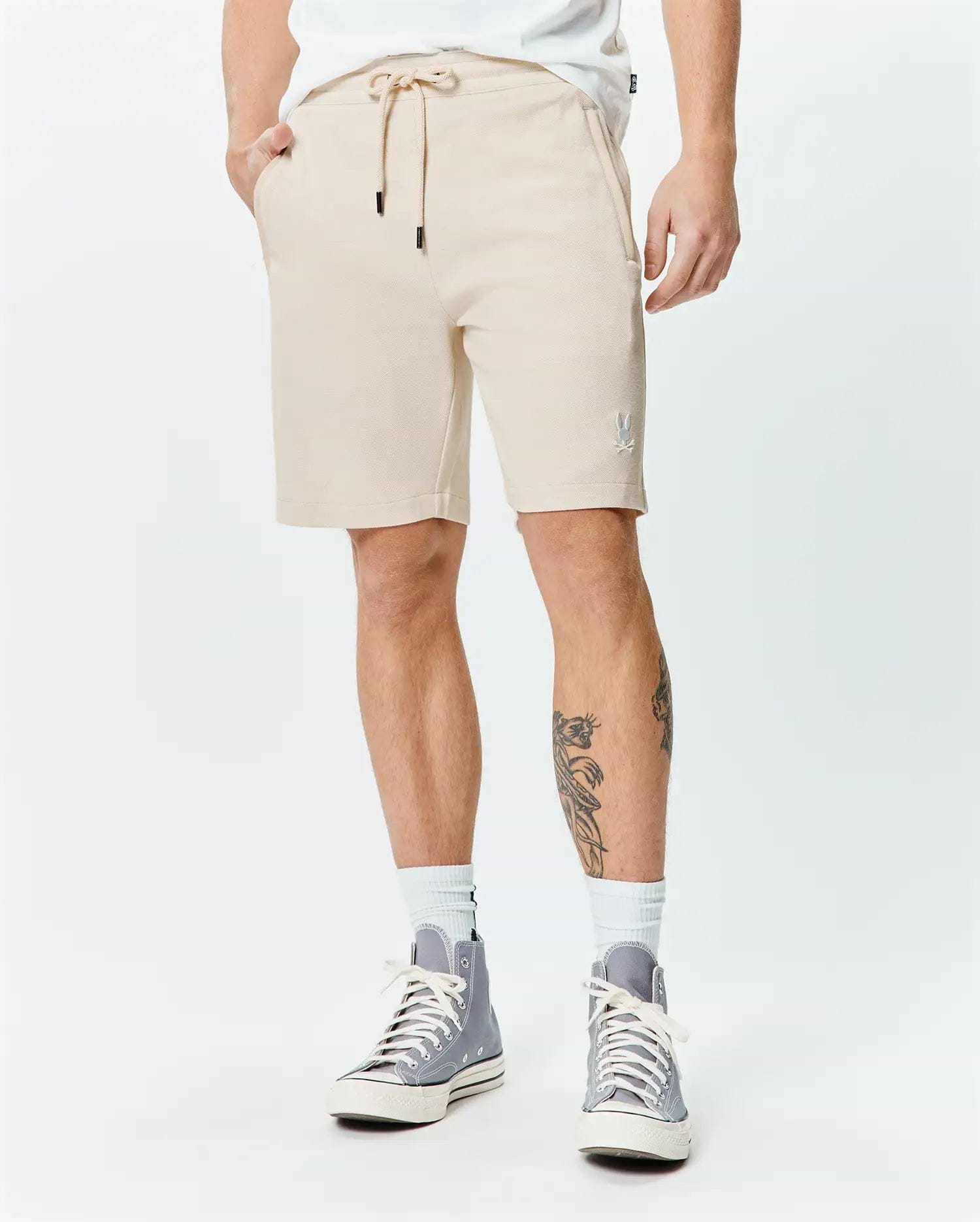 Mens Keith Short 8, sand pique knit, relaxed fit shorts with drawstring waist, black tips, and a small white embroidered Psycho Bunny skull logo on the left thigh.