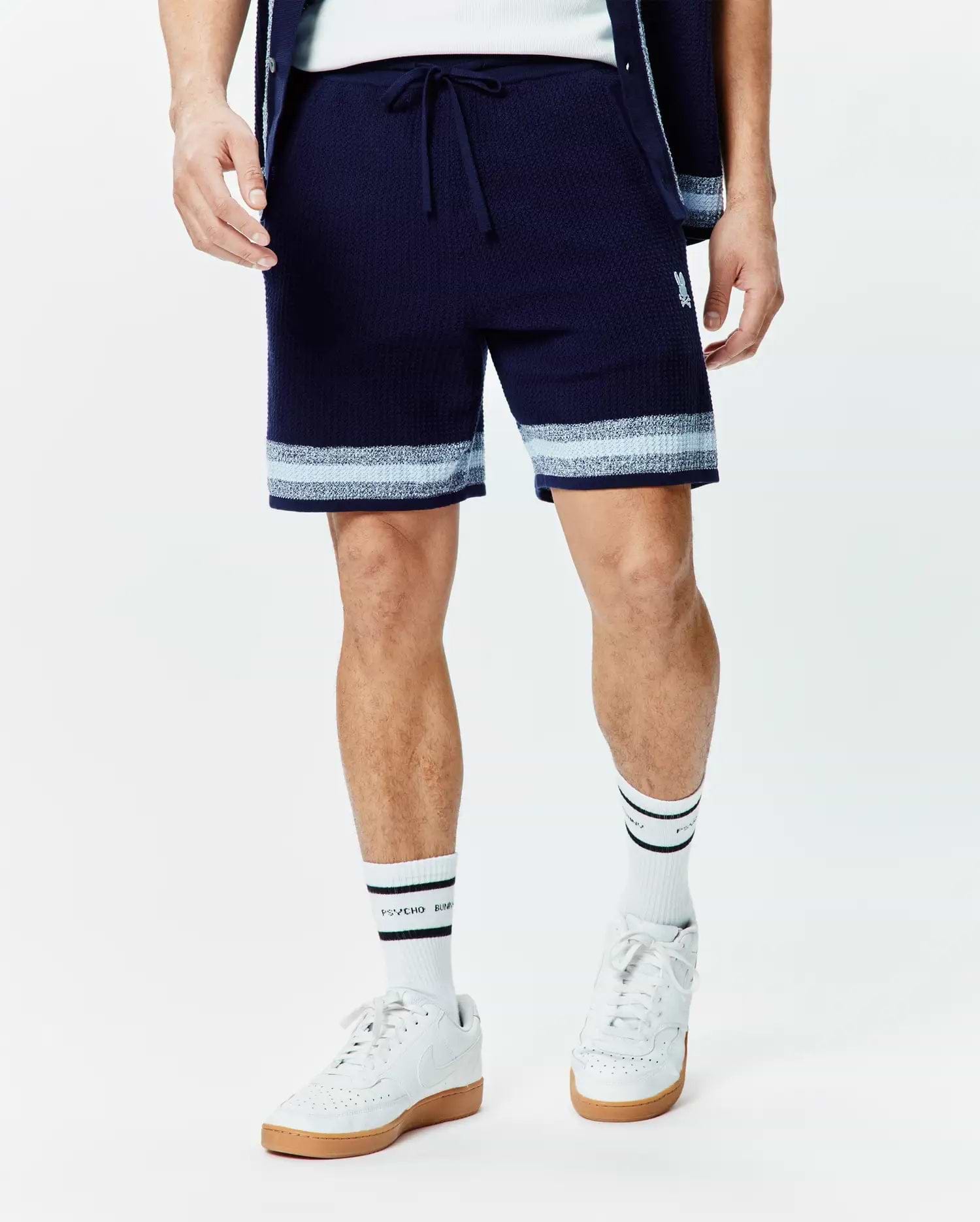 Mens Giles Knit Short 7, a navy textured knit with a drawstring waist, featuring light blue and white horizontal stripes at the hem and a small embroidered white Psycho Bunny skull logo on the left thigh for a relaxed fit.
