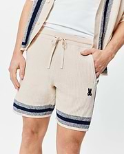 Mens Giles Knit Short 7 in cream textured knit, featuring a drawstring waist, side pockets, navy and light blue striped hems, and a small embroidered Psycho Bunny skull logo on the left leg.