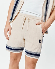 Mens Giles Knit Short 7 in cream textured knit, featuring a drawstring waist, side pockets, navy and light blue striped hems, and a small embroidered Psycho Bunny skull logo on the left leg.