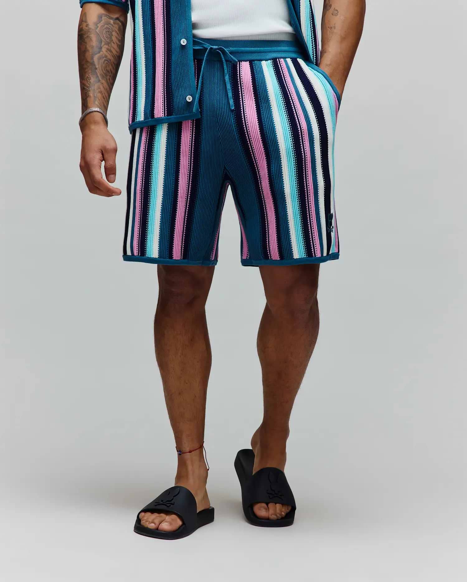 Mens Cliff Knit Short, a relaxed fit knit short with bold teal, pink, white, light blue, and navy vertical stripes, featuring a small embroidered Psycho Bunny skull logo on the lower left leg and a drawstring waist. (3)