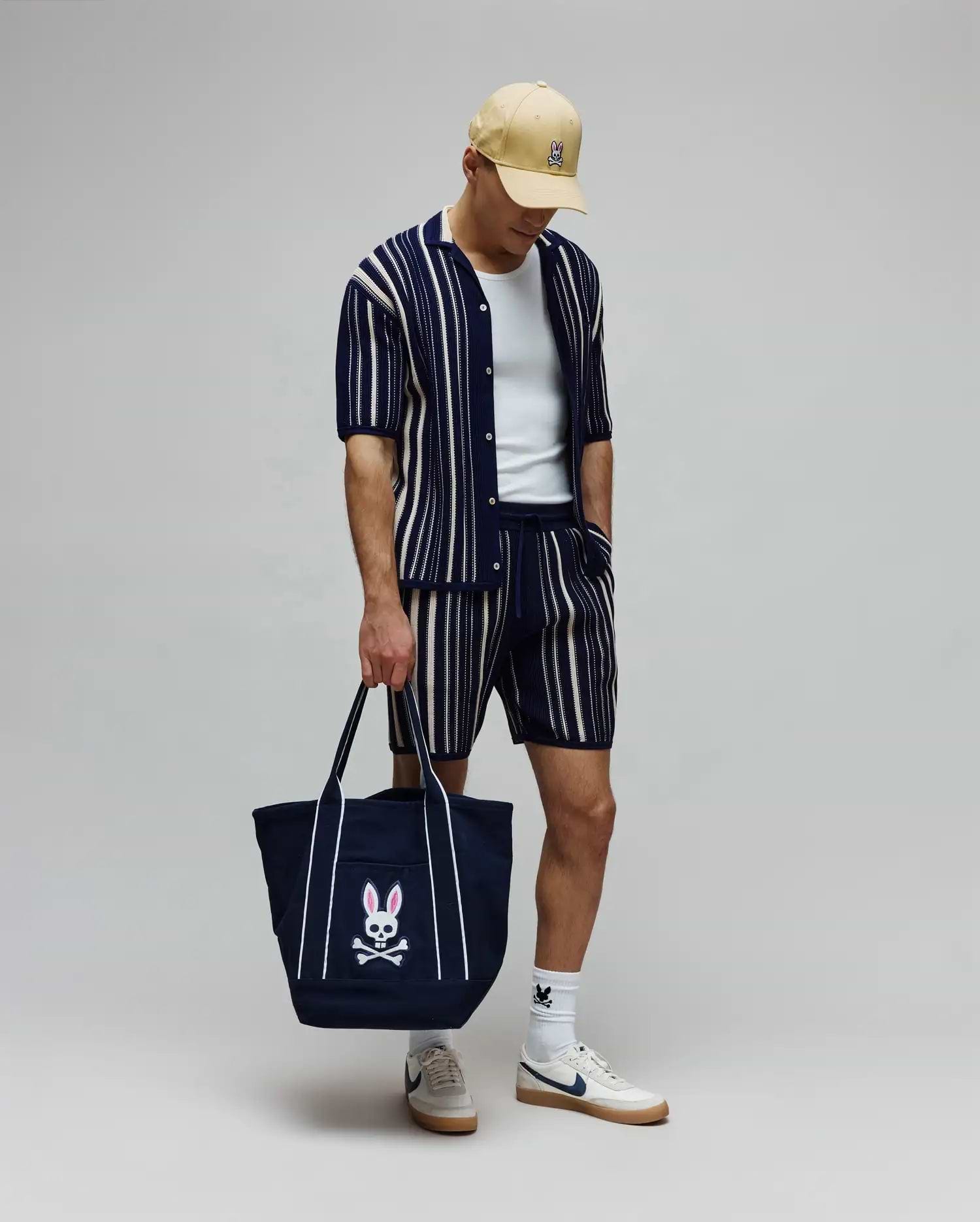 Mens Cliff Knit Short and matching knit polo, both navy with cream vertical stripes, a tan cap, white socks, and a navy tote bag, all featuring the iconic embroidered Psycho Bunny skull logo. (3)