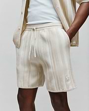 Mens Cliff Knit Short in cream with subtle vertical beige ribbed texture, featuring a relaxed fit, drawstring waist, and a small embroidered Psycho Bunny skull logo on the lower right leg.