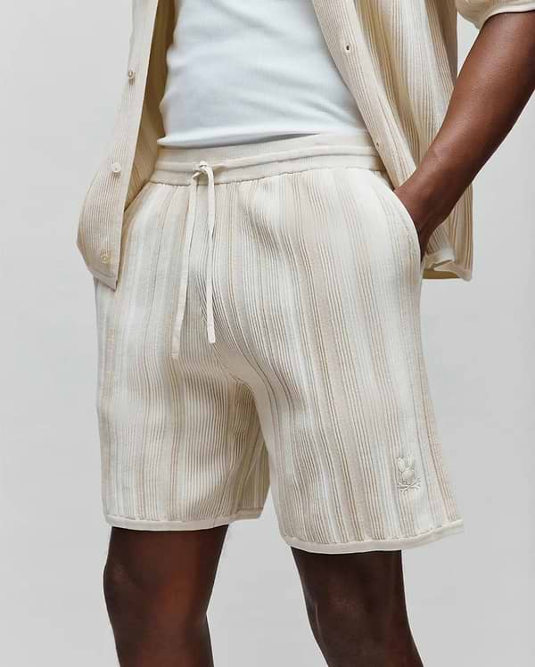 Mens Cliff Knit Short in cream with subtle vertical beige ribbed texture, featuring a relaxed fit, drawstring waist, and a small embroidered Psycho Bunny skull logo on the lower right leg.