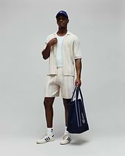 Mens Cliff Knit Short in cream, a relaxed fit with vertical ribbed knit texture and a small embroidered Psycho Bunny skull logo on the left thigh. Complemented by a matching ribbed shirt and a navy Psycho Bunny tote.