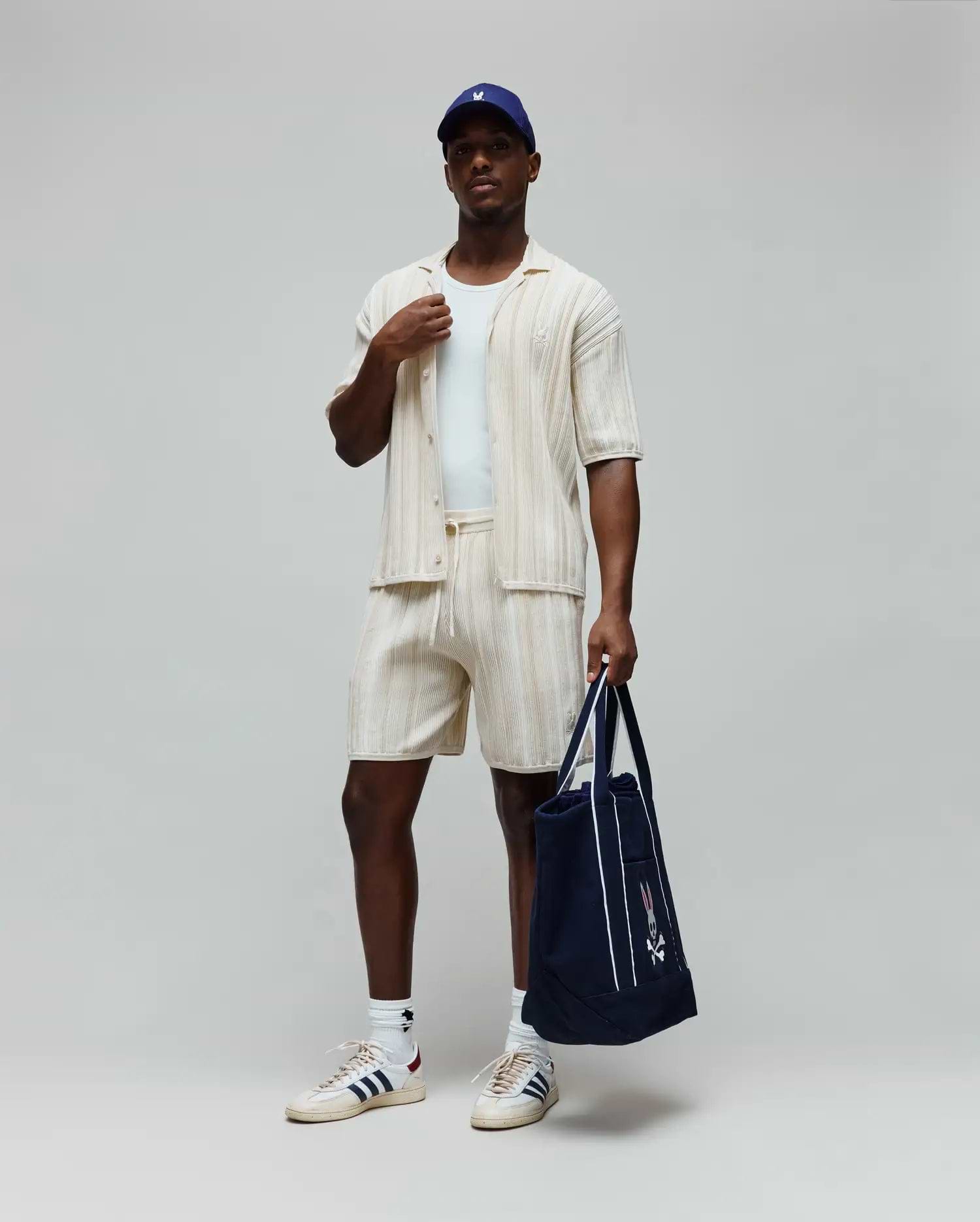 Mens Cliff Knit Short in cream, a relaxed fit with vertical ribbed knit texture and a small embroidered Psycho Bunny skull logo on the left thigh. Complemented by a matching ribbed shirt and a navy Psycho Bunny tote.