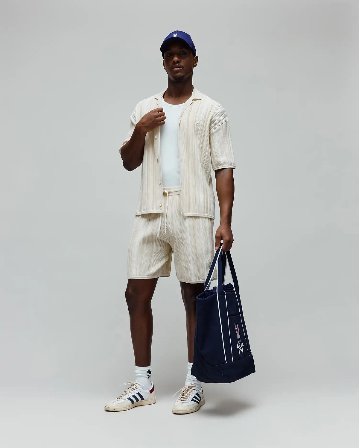 Mens Cliff Knit Short in cream, a relaxed fit with vertical ribbed knit texture and a small embroidered Psycho Bunny skull logo on the left thigh. Complemented by a matching ribbed shirt and a navy Psycho Bunny tote.