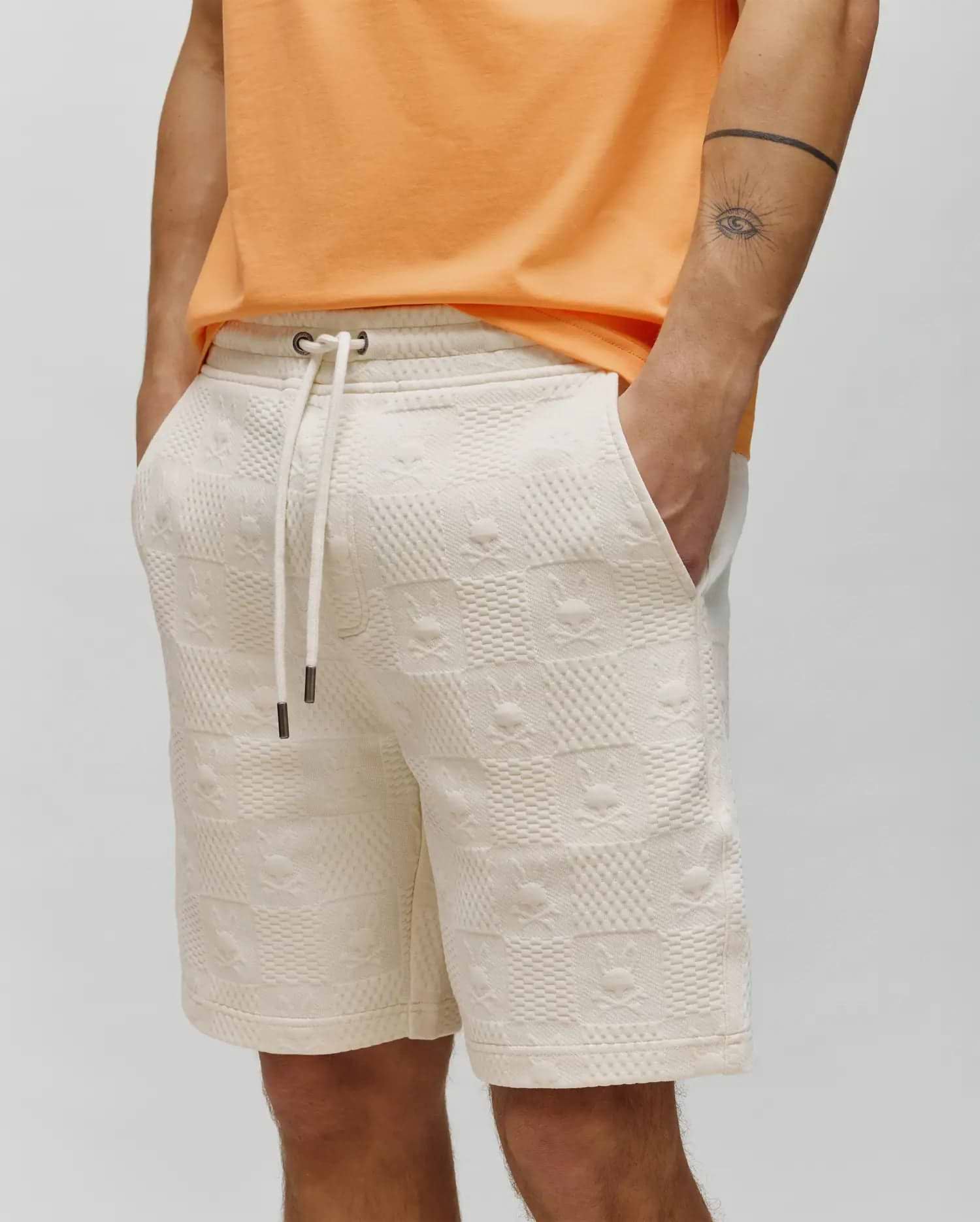 Cream Mens Dustin Sweatshort with a relaxed fit, featuring an all-over embossed Psycho Bunny skull and crossbones pattern on textured French terry fabric, and a drawstring waist with metal tips.