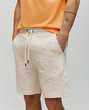 Cream Mens Dustin Sweatshort with a relaxed fit, featuring an all-over embossed Psycho Bunny skull and crossbones pattern on textured French terry fabric, and a drawstring waist with metal tips.
