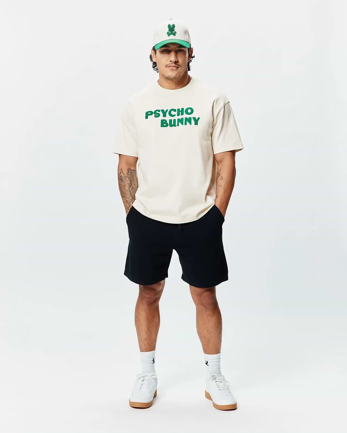 Cream classic fit jersey t-shirt with bold green PSYCHO BUNNY chest text, paired with black Mens Devon Knit Short, a cream and green Psycho Bunny skull logo cap, and white logo socks.