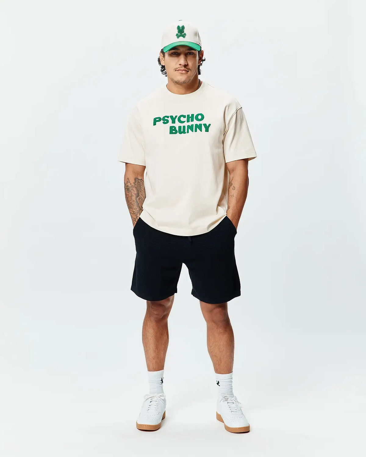 Cream classic fit jersey t-shirt with bold green PSYCHO BUNNY chest text, paired with black Mens Devon Knit Short, a cream and green Psycho Bunny skull logo cap, and white logo socks.
