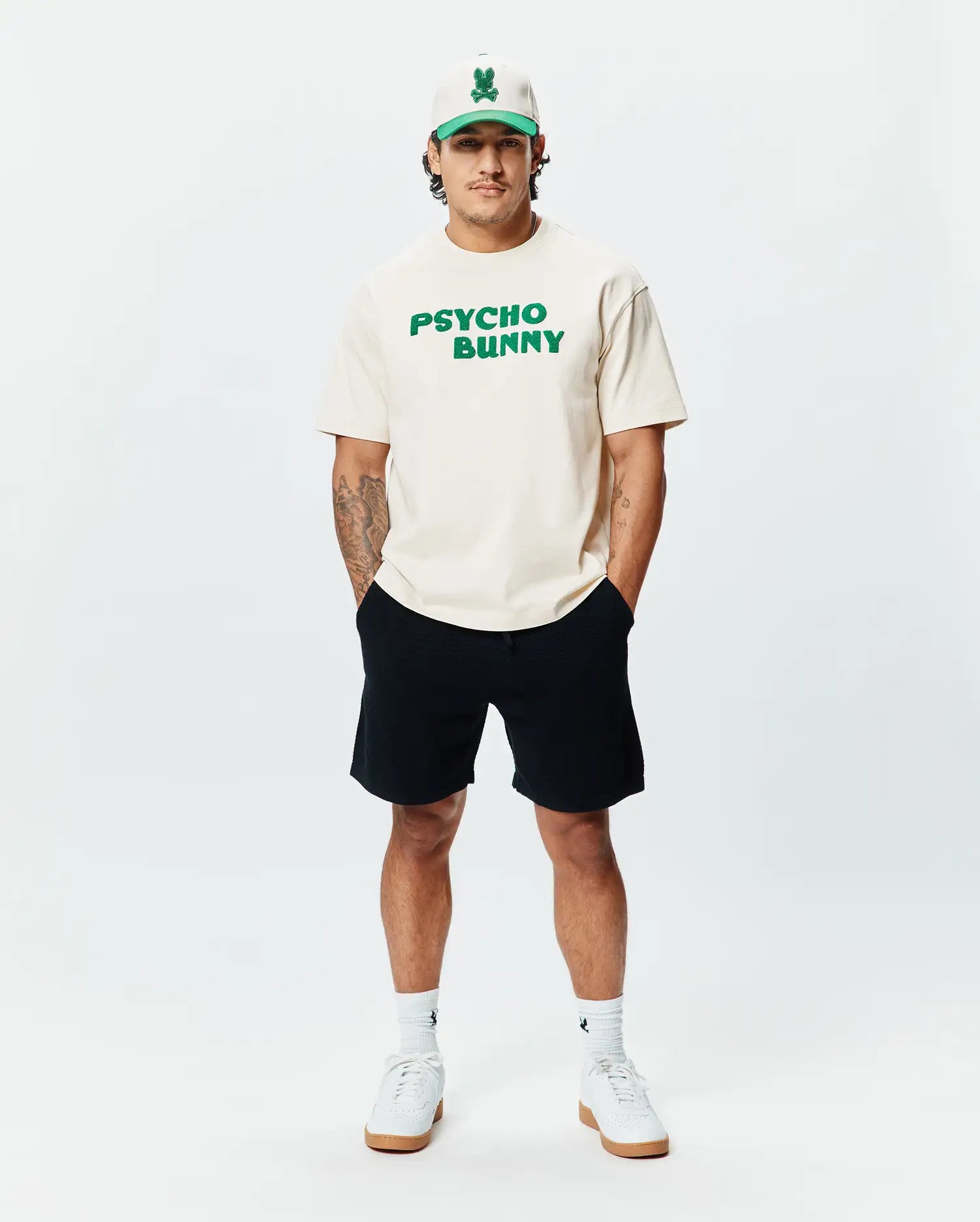 Cream classic fit jersey t-shirt with bold green PSYCHO BUNNY chest text, paired with black Mens Devon Knit Short, a cream and green Psycho Bunny skull logo cap, and white logo socks.