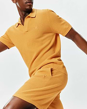 Golden yellow textured knit polo shirt with ribbed collar and cuffs, paired with matching Mens Devon Knit Short, both featuring a small embroidered Psycho Bunny skull logo for a classic fit.