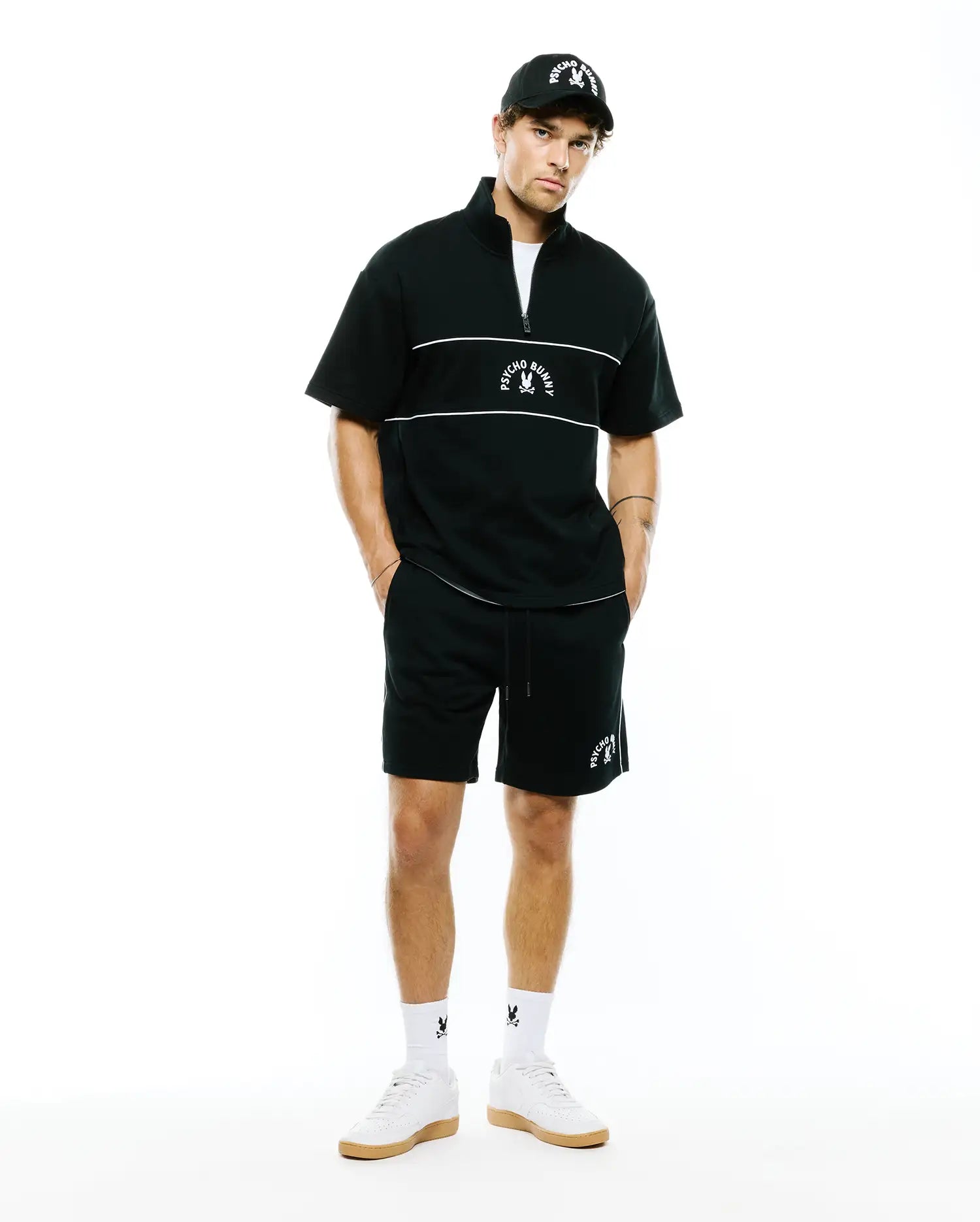 Black short-sleeve quarter-zip top with white chest stripes and embroidered Psycho Bunny logo, paired with MENS STEVIE SHORT featuring white side piping and vertical logo. Includes logo cap and socks.