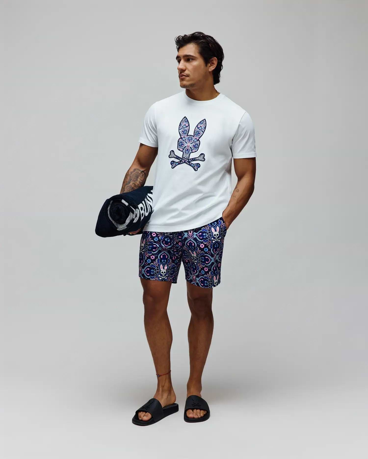 White classic fit jersey t-shirt with a large, colorful paisley Psycho Bunny skull logo on the chest, and Mens Luke Swim Trunk in navy with an all-over intricate pattern featuring small bunny skulls.