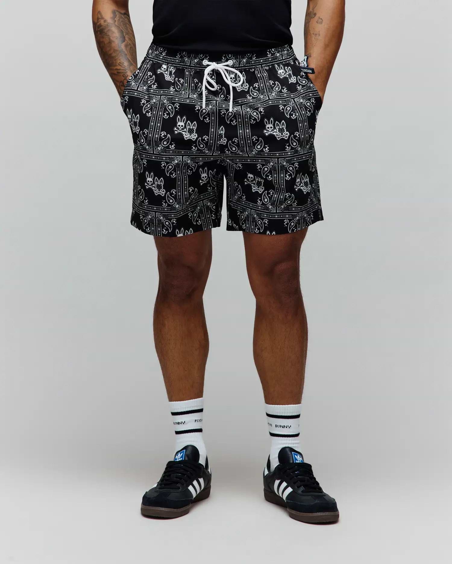 Mens Luke Swim Trunk in black with a white all-over bandana paisley print featuring integrated Psycho Bunny skull logos, a relaxed fit, and a white drawstring.