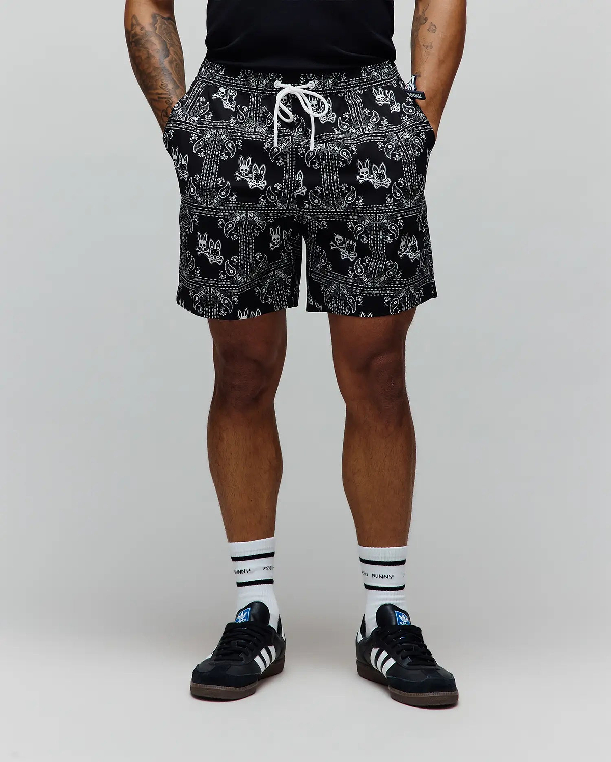 Mens Luke Swim Trunk in black with a white all-over bandana paisley print featuring integrated Psycho Bunny skull logos, a relaxed fit, and a white drawstring.