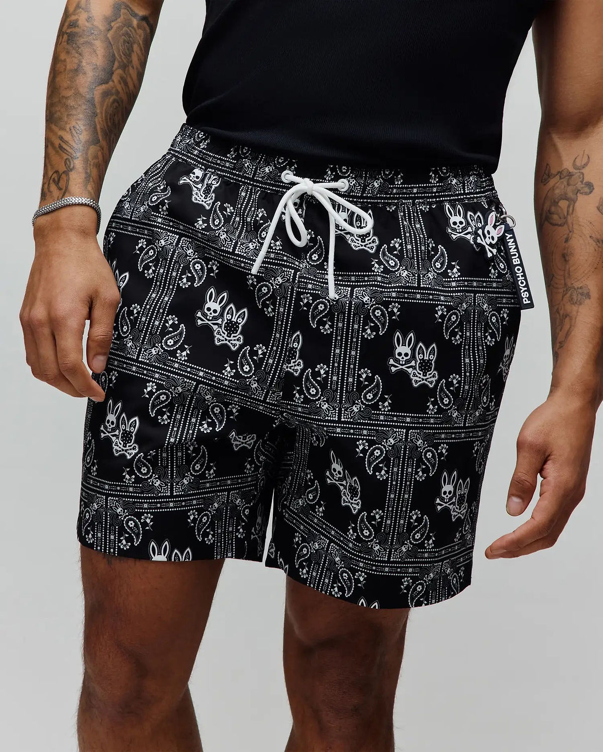 Mens Luke Swim Trunk, black with an all-over white paisley bandana print featuring the iconic Psycho Bunny skull logo, white drawstring, and a branded Psycho Bunny tag.