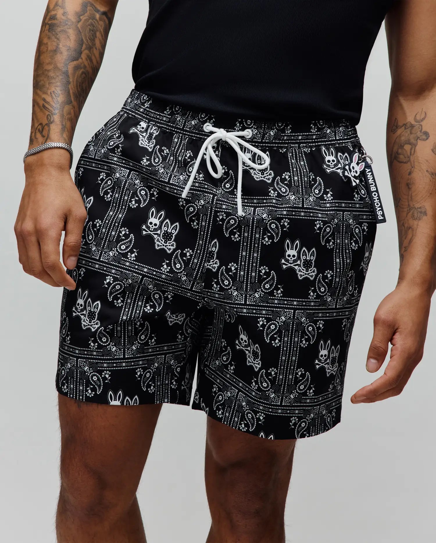 Mens Luke Swim Trunk, black with an all-over white paisley bandana print featuring the iconic Psycho Bunny skull logo, white drawstring, and a branded Psycho Bunny tag.