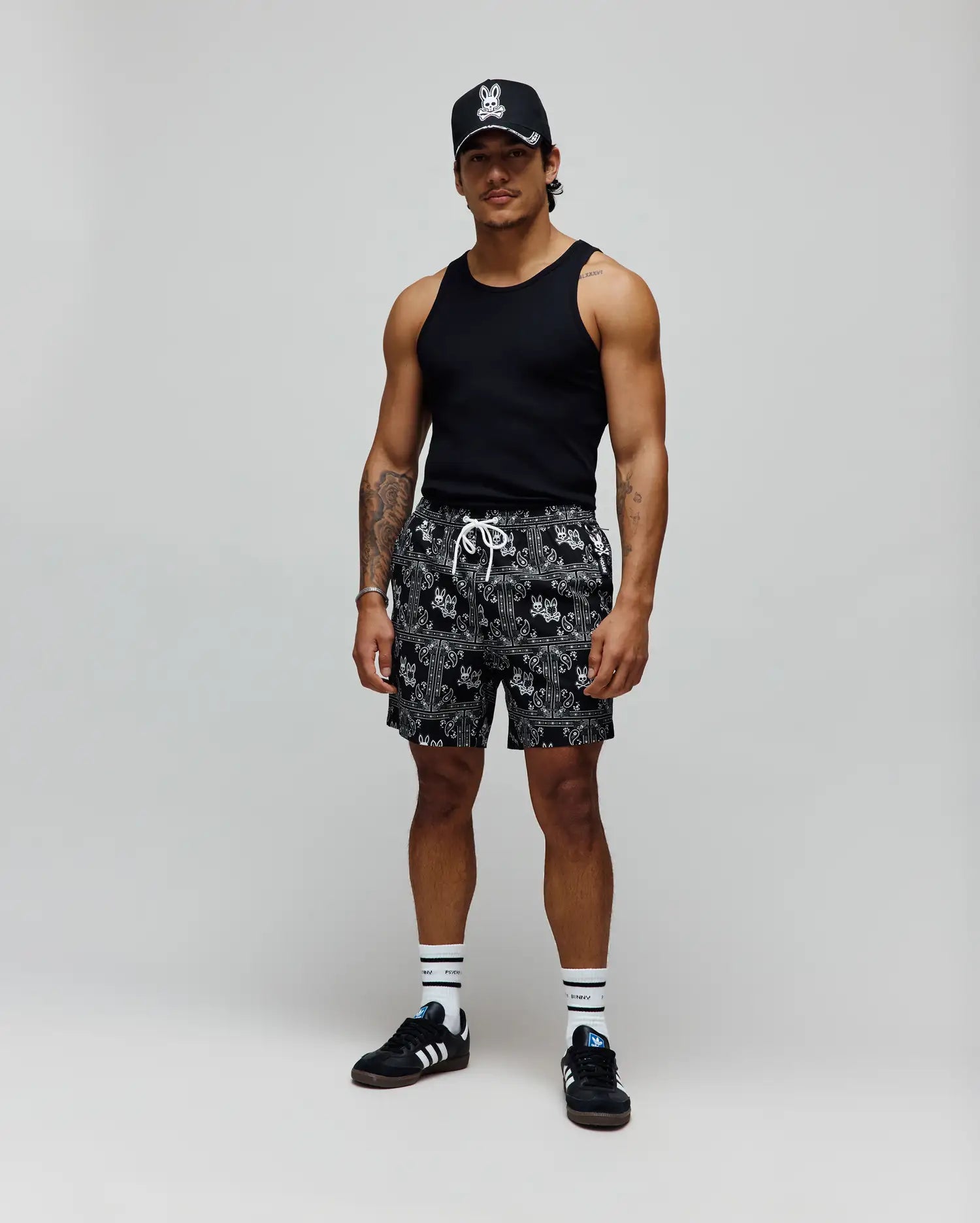 Mens Luke Swim Trunk in black with white bandana print featuring repeated small Psycho Bunny skull logos and white drawstring. Worn with a black tank top and black cap with large white Psycho Bunny logo.
