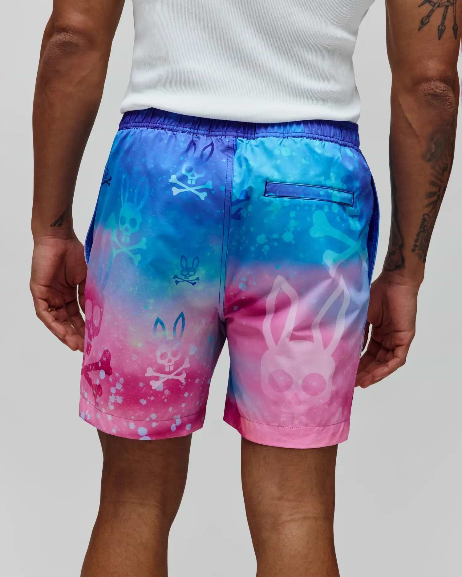 Mens Caspian Swim Trunk, vibrant blue-to-pink galaxy gradient with all-over Psycho Bunny skull and crossbones print, relaxed fit, smooth quick-dry fabric, and purple elastic waistband.