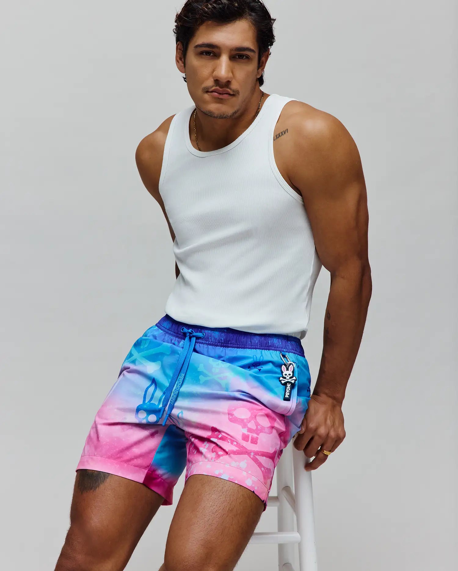 Mens Caspian Swim Trunk with a vibrant blue to pink tie-dye gradient, subtle skull pattern, dark blue waistband, and a Psycho Bunny logo charm on the drawstring with vertical branding.