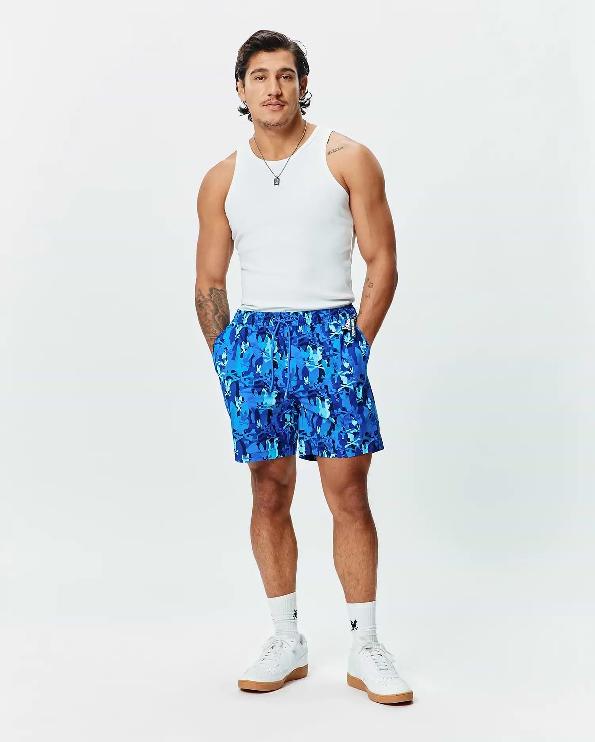 Mens Tanner Swim Trunk, a relaxed fit blue swim trunk featuring an all-over camouflage print composed of repeating Psycho Bunny skull logos, with a drawstring waist and a small brand tag.