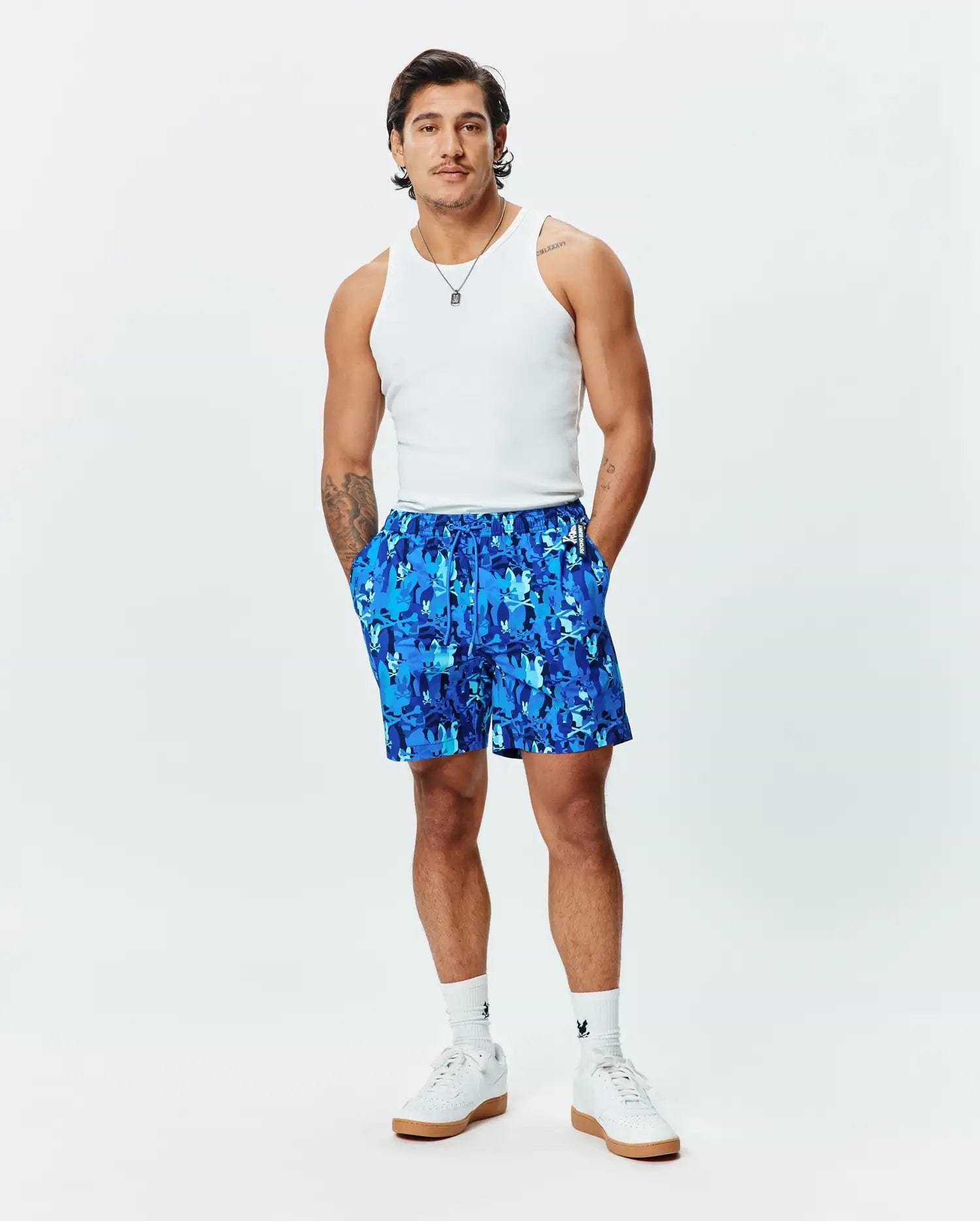 Mens Tanner Swim Trunk, a relaxed fit blue swim trunk featuring an all-over camouflage print composed of repeating Psycho Bunny skull logos, with a drawstring waist and a small brand tag.