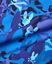Tanner Swim Trunk in a bold blue and purple abstract camouflage print, featuring the iconic Psycho Bunny skull and crossbones logo subtly integrated throughout the smooth, quick-dry fabric.