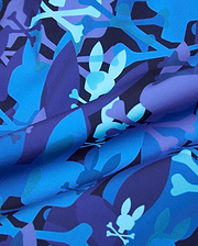 Tanner Swim Trunk in a bold blue and purple abstract camouflage print, featuring the iconic Psycho Bunny skull and crossbones logo subtly integrated throughout the smooth, quick-dry fabric.