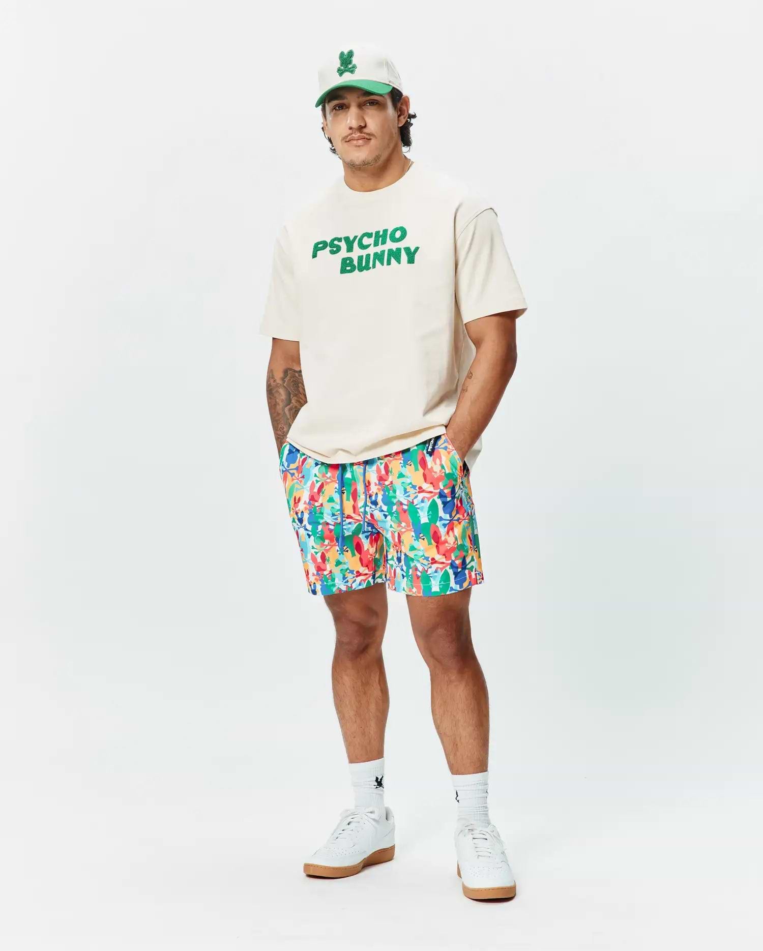 Relaxed fit cream jersey t-shirt with bold green PSYCHO BUNNY chest graphic, vibrant multi-color abstract floral Mens Tanner Swim Trunk, white cap with green bunny skull logo, and white logo socks.
