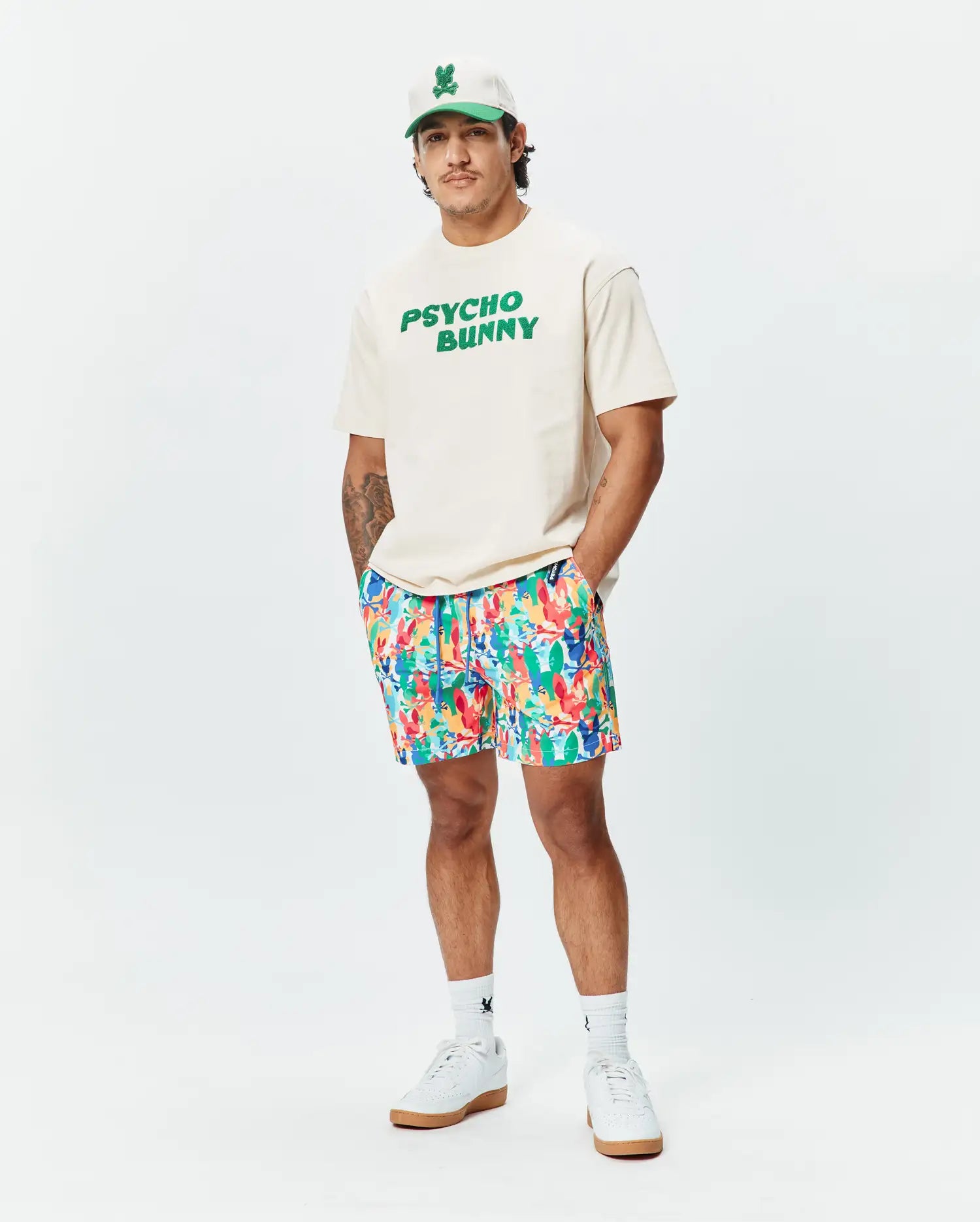 Relaxed fit cream jersey t-shirt with bold green PSYCHO BUNNY chest graphic, vibrant multi-color abstract floral Mens Tanner Swim Trunk, white cap with green bunny skull logo, and white logo socks.