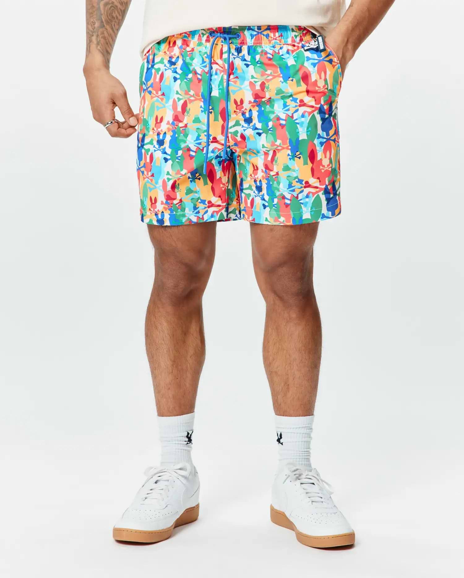 Mens Tanner Swim Trunk featuring a vibrant all-over print of colorful Psycho Bunny skull logos, crossbones, and abstract shapes, with a blue drawstring and a classic fit.