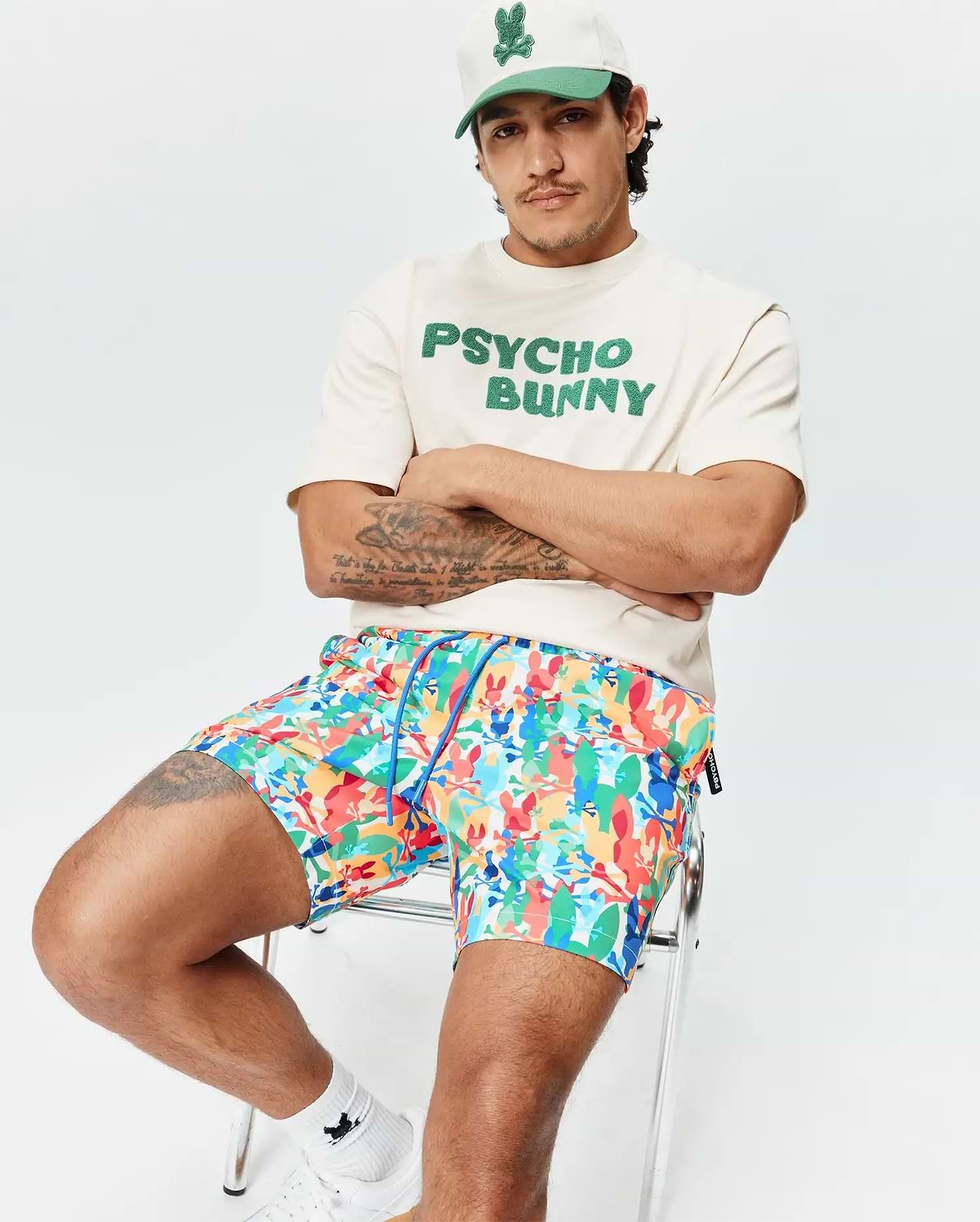 Mens Tanner Swim Trunk in vibrant multi-color with all-over Psycho Bunny skull print and blue drawstring. Styled with a cream jersey t-shirt featuring bold green PSYCHO BUNNY text and a two-tone cap with bunny logo.