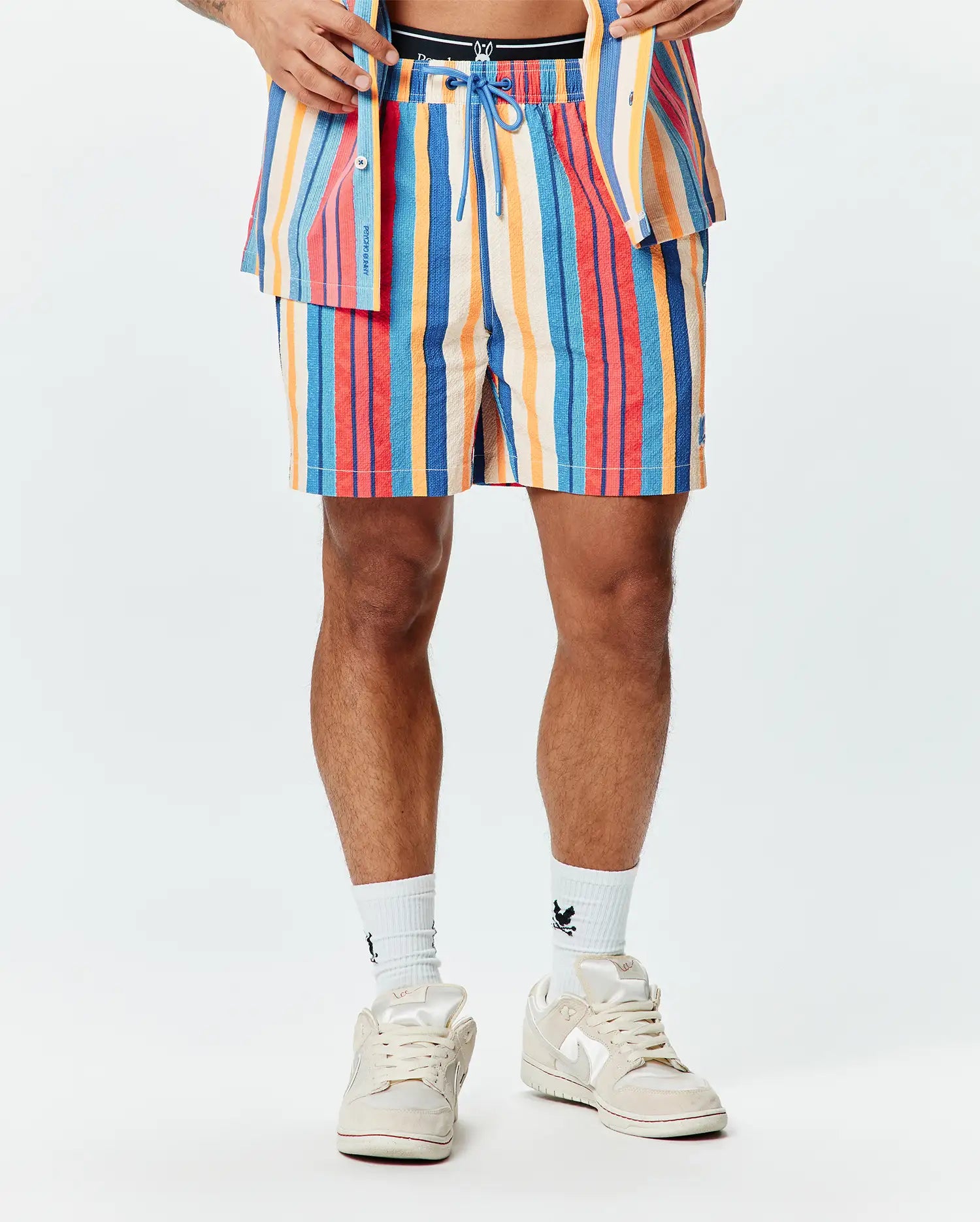 Mens Hubert Swim Trunk, a multi-colored striped swim trunk with blue, red, orange, and cream vertical stripes, featuring a textured fabric, blue drawstring, and a small embroidered Psycho Bunny skull logo on the left leg.