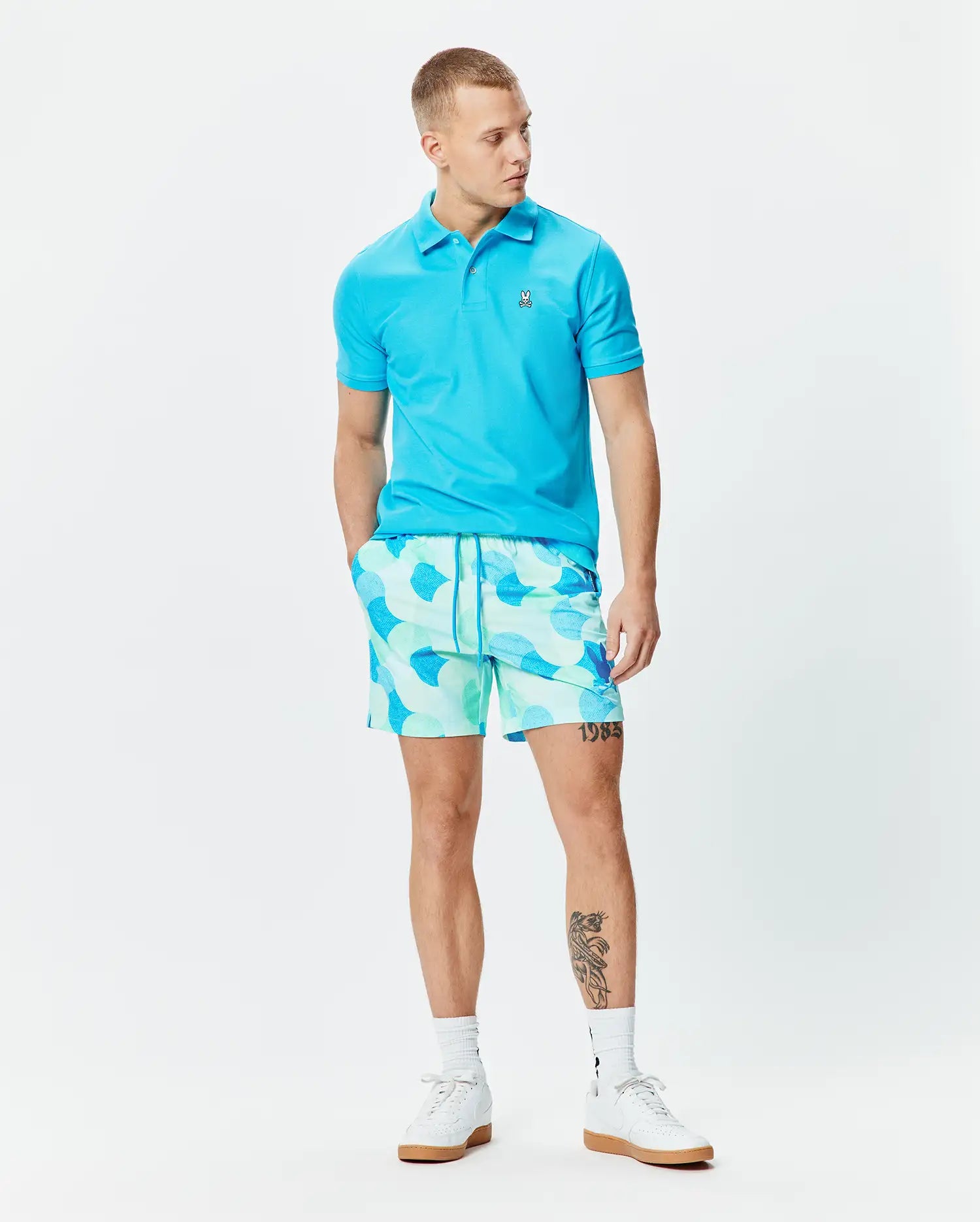 Bright blue pique polo with small embroidered Psycho Bunny skull logo on chest, paired with Mens Ethan Swim Trunk 6 featuring a light mint and blue abstract pattern and drawstring waist.