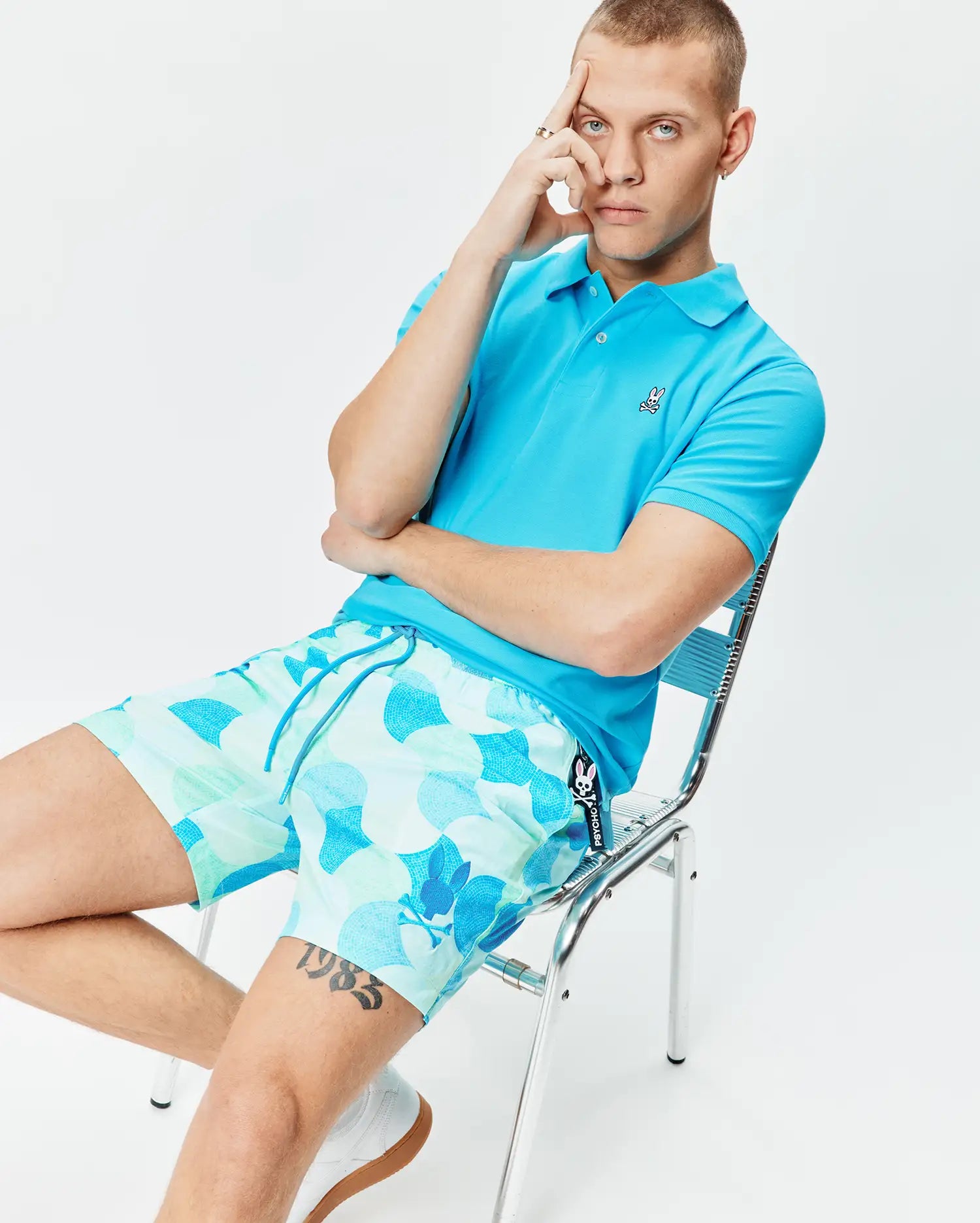 Bright blue pique polo with embroidered Psycho Bunny logo on chest, paired with Mens Ethan Swim Trunk 6 featuring an aqua and blue wavy geometric pattern and side Psycho Bunny tag.