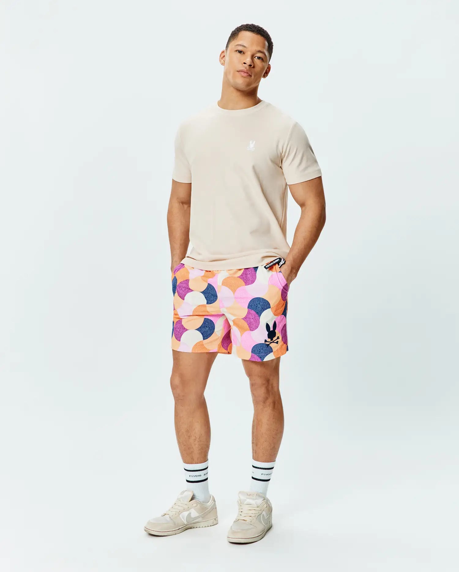 Beige classic fit jersey t-shirt with small white embroidered Psycho Bunny logo on left chest, paired with Mens Ethan Swim Trunk 6 featuring a vibrant abstract pink, orange, purple, blue wavy pattern and a large black Psycho Bunny logo on the left leg.