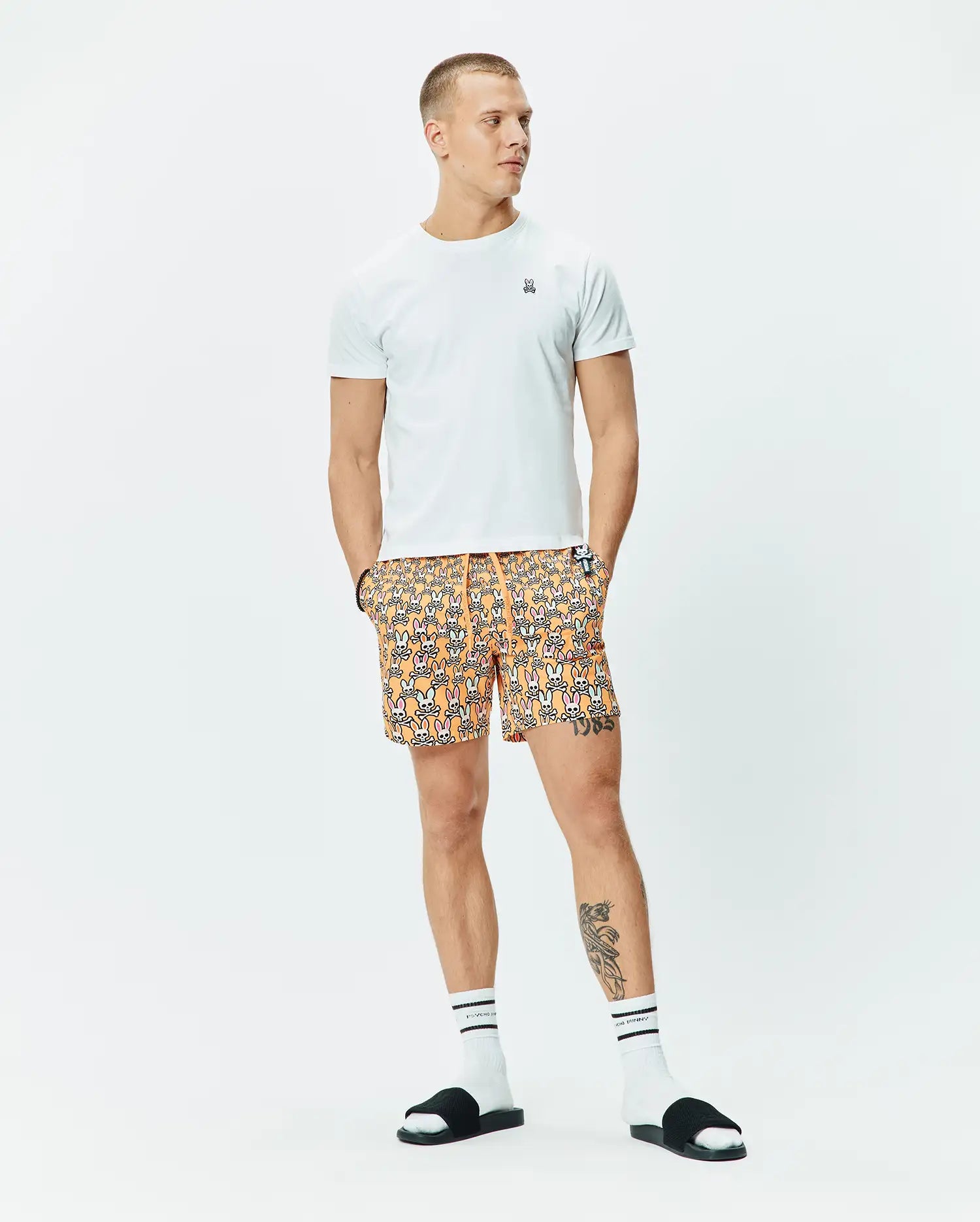 White t-shirt with small embroidered Psycho Bunny logo, complementing Mens Byron Swim Trunk 6, an orange all-over print with repeating Psycho Bunny skulls, and white striped Psycho Bunny socks.