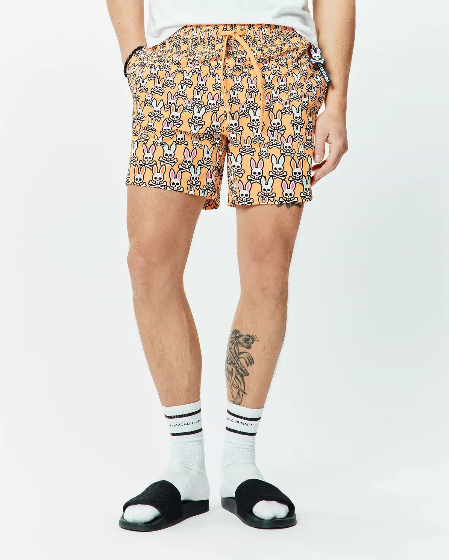 Mens Byron Swim Trunk 6, orange with all-over pink, light blue, white, and black Psycho Bunny skull pattern, classic fit, smooth fabric, small side logo tag.